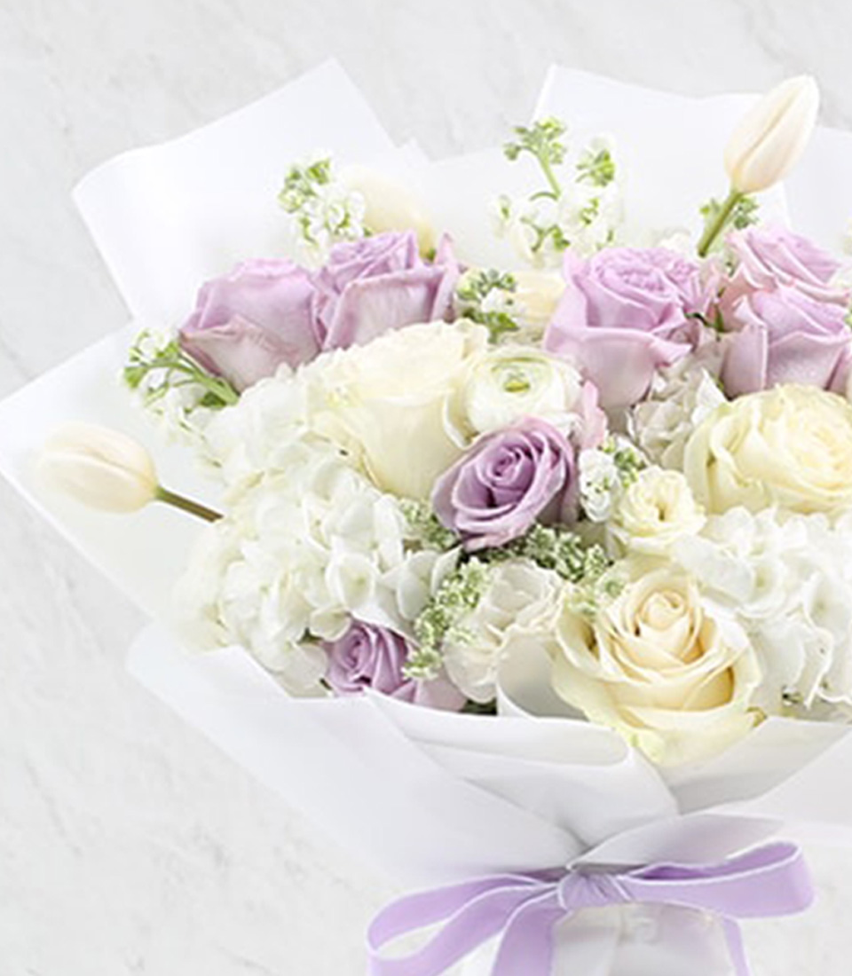 A stunning bouquet of white, lavender, and pale pink roses ensconced in luxurious white wrapping paper with a delicate lavender ribbon bearing the Basketeer logo graces the marble backdrop. Nestled beside this fragrant floral arrangement is a bottle of fine prosecco, exuding elegance and celebration. A lavish gift card inscribed with 