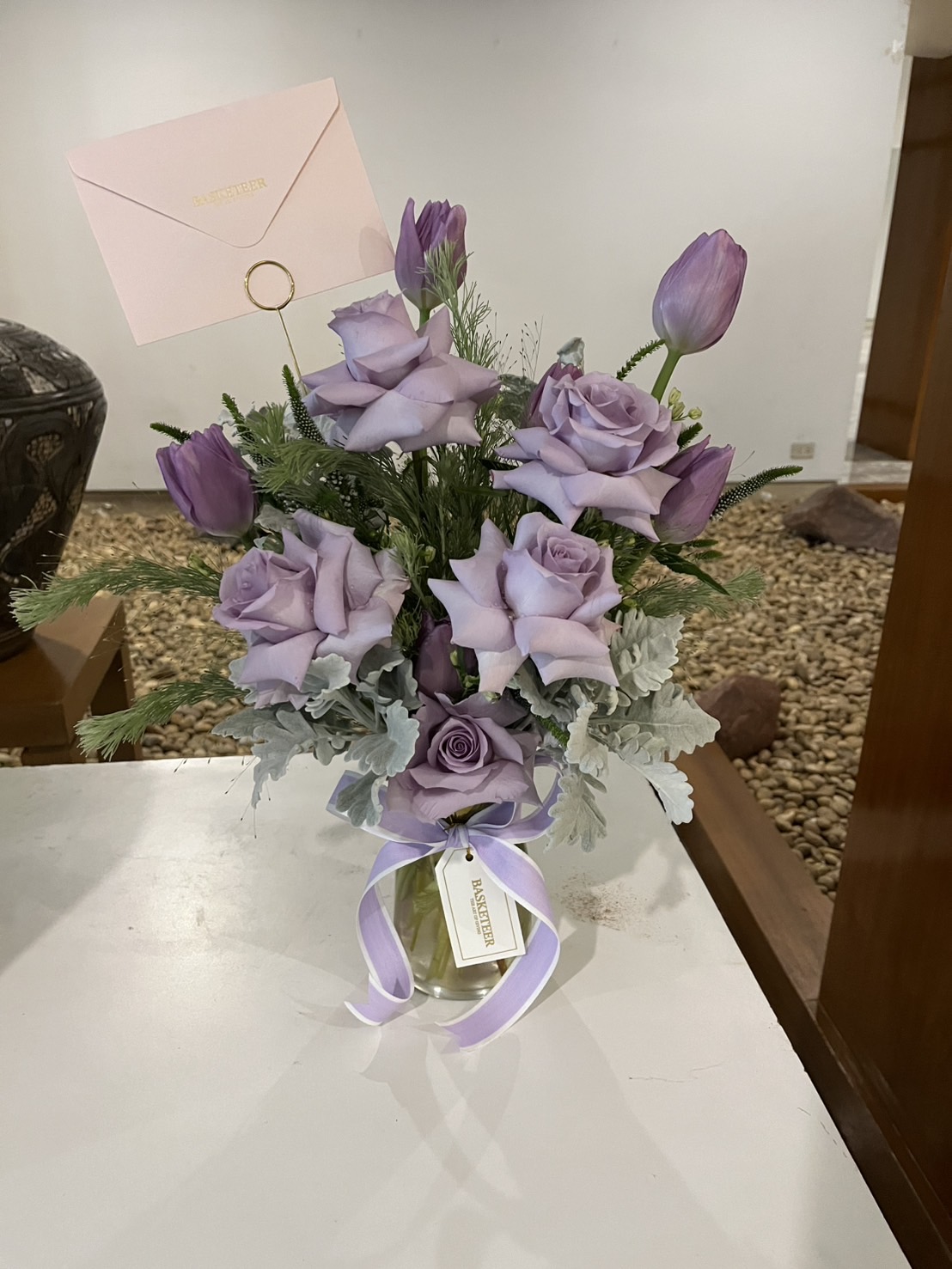 Elegant glass vase with lavender roses, purple tulips, dusty miller leaves, and greenery, tied with a lavender satin ribbon.