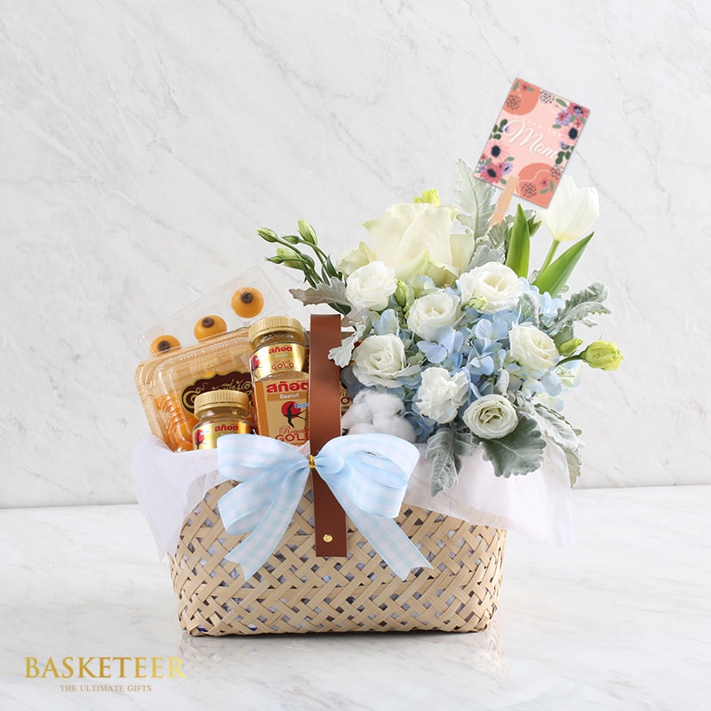 A woven gift basket, adorned with a blue and white ribbon, contains white and blue flowers, packaged snacks, and a small floral greeting card. The basket is placed on a marble surface, with the brand 