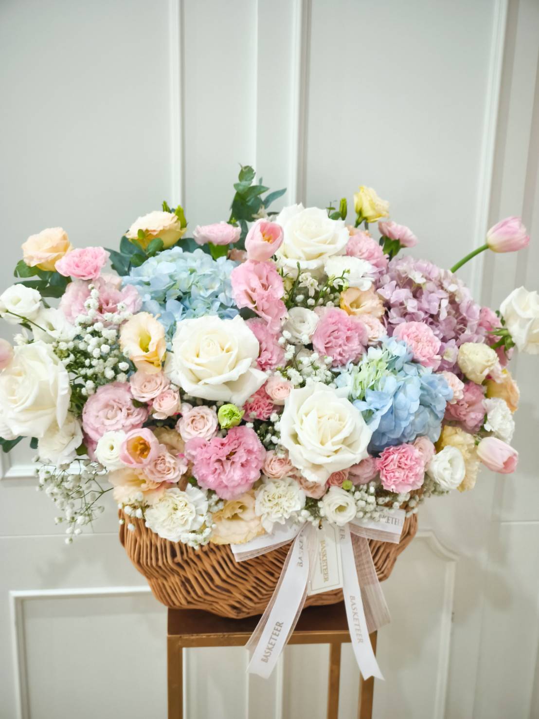 Pastel flower basket with roses, hydrangeas, tulips, lisianthus, and peonies in soft pink, white, blue, and yellow tones.