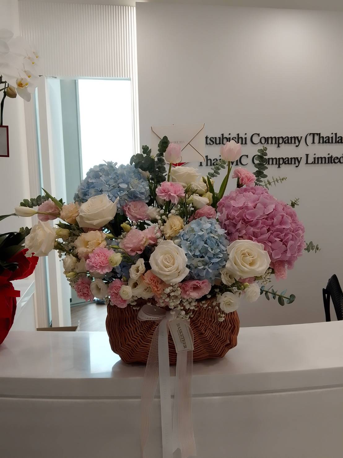A Gentle Sweet Hues Flowers Basket filled with blue, pink, and white hydrangeas, roses, and carnations sits on a white reception desk in an office with 