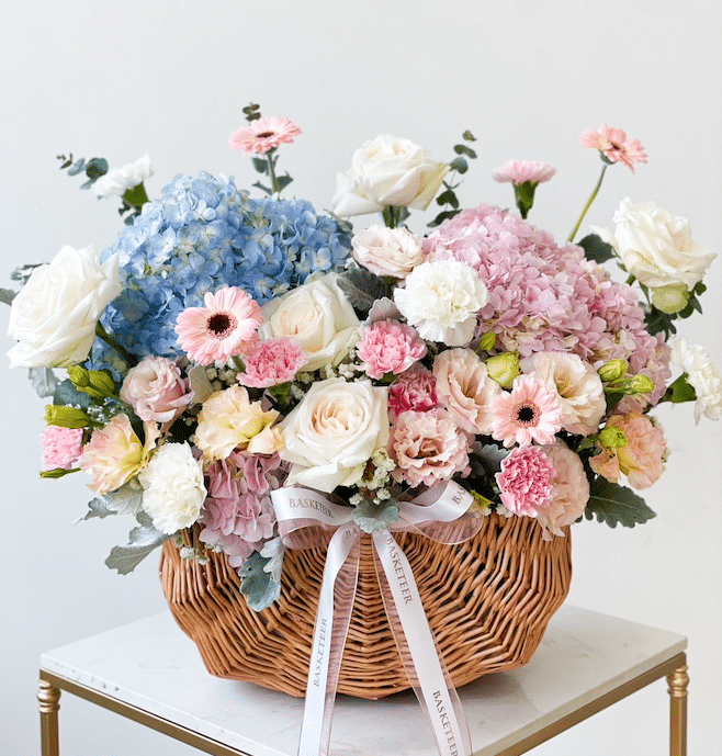 A wicker basket filled with a variety of flowers, including blue and pink hydrangeas, white roses, pink carnations, and white and peach gerberas. The basket is adorned with white ribbons, and the arrangement sits on a white table against a plain background.