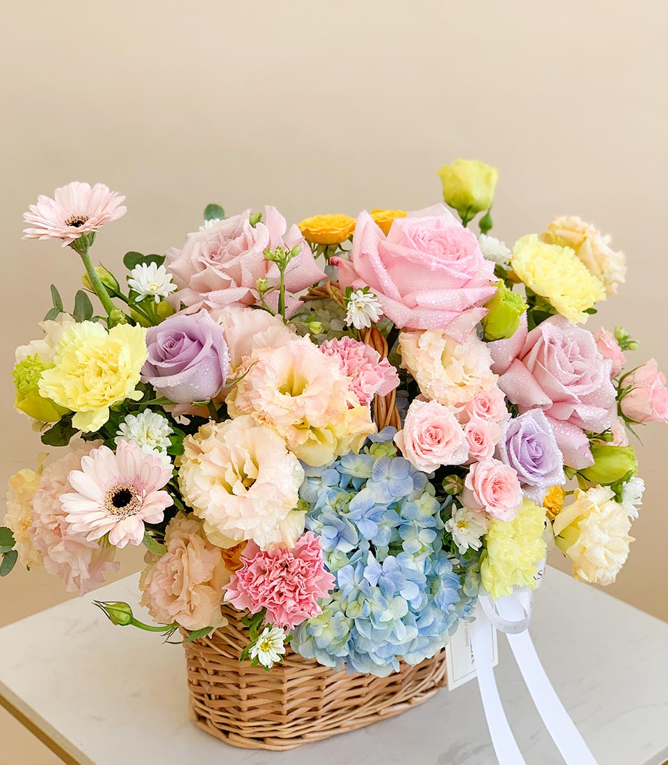 Pastel flower basket with pink roses, purple hydrangeas, and assorted fresh blooms in a woven wicker basket.