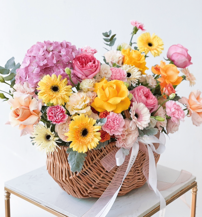 A wicker basket brimming with a vibrant assortment of flowers including yellow and white daisies, pink and orange roses, pink carnations, purple hydrangeas, and greenery. The basket is adorned with a white ribbon and is placed on a small table.