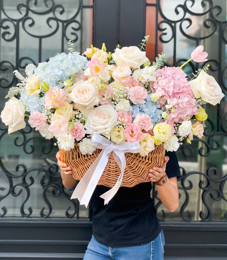 Person holding a large wicker basket with a white ribbon, filled with a colorful arrangement of flowers including white roses, pink and blue hydrangeas, and various pastel blooms, in front of an ornate black metal gate.
