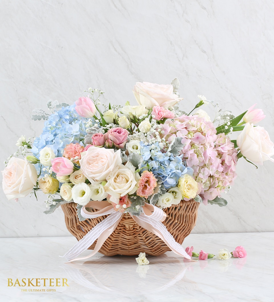 A sumptuous wicker basket brimming with a luxurious array of florals, featuring pristine white roses, delicate pink tulips, and vibrant blue and pink hydrangeas intertwined with lush greenery. The basket is elegantly adorned with a crisp white ribbon bow that exudes sophistication. Resting on a refined light marble surface, the opulent setup is enhanced by the prominent 