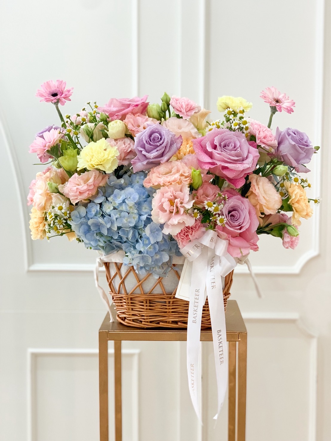 Pastel floral basket arrangement with purple, pink, and cream roses, hydrangeas, carnations, and lisianthus in a rattan basket with white ribbon.