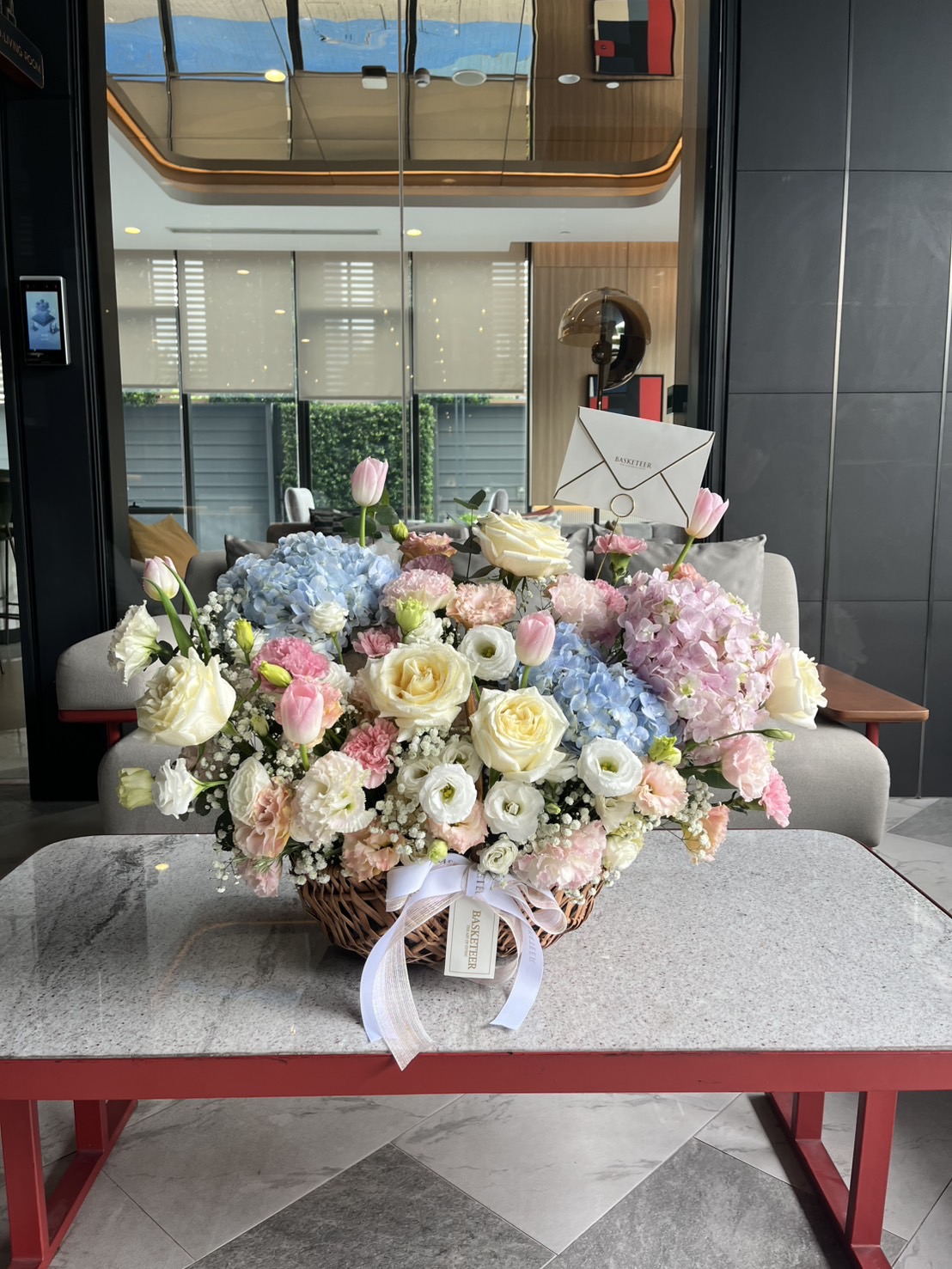 A Sweet Flowers Basket with pastel roses, hydrangeas, and tulips sits on a marble coffee table in a modern living room. An envelope and white ribbon are attached—perfect for Mother’s Day or paired with a sky blue cake.