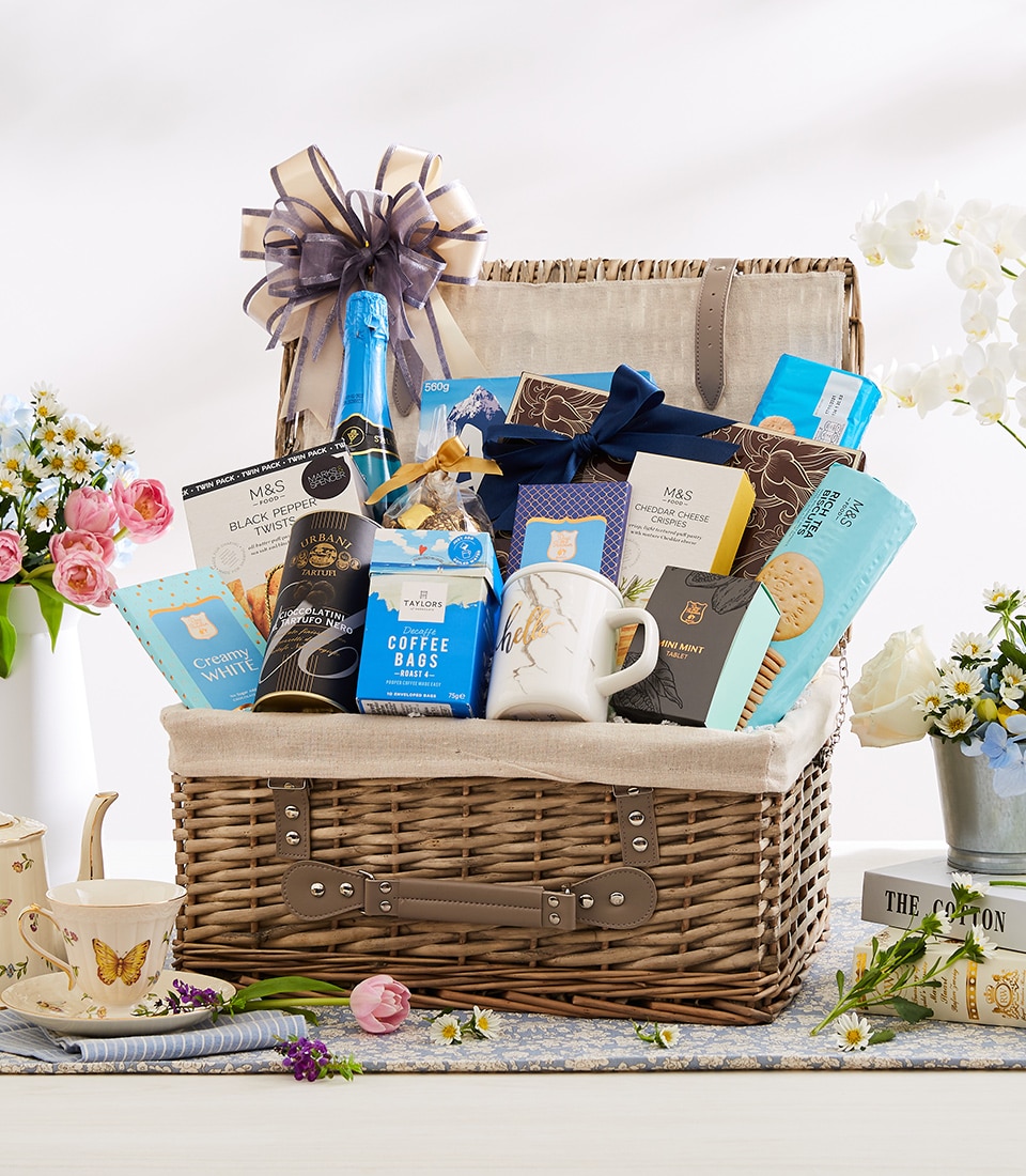 A luxurious wicker basket brimming with an exquisite selection of gift items, including gourmet tea, decadent chocolates, artisan cookies, premium coffee bags, a chic blue bottle, and a pristine white mug. This elegant ensemble is adorned with a sumptuous ribbon featuring the Basketeer logo and is artfully surrounded by vibrant flowers, creating an inviting and sophisticated presentation perfect for any opulent occasion.