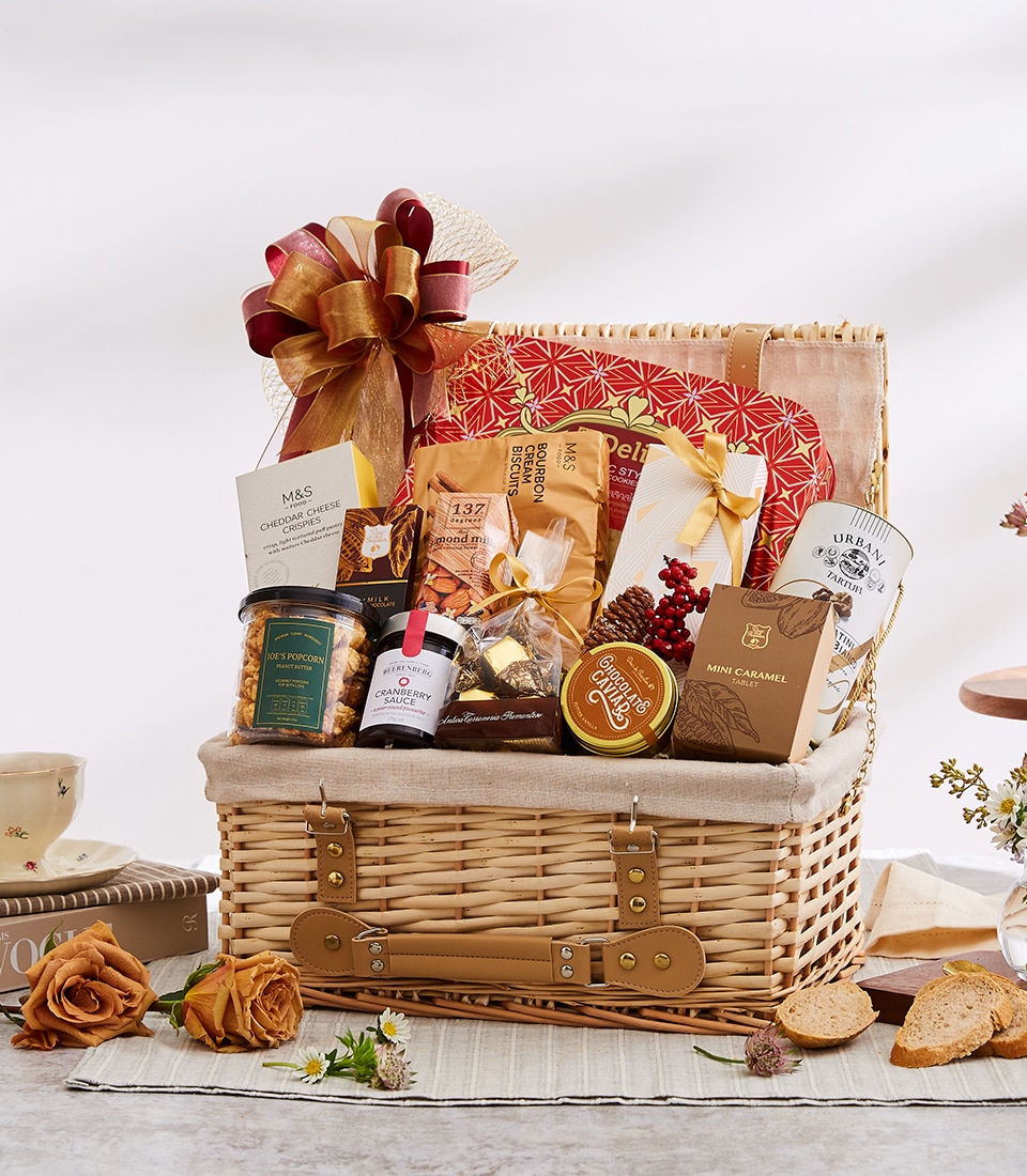 A luxurious wicker gift basket by Basketeer, brimming with an exquisite selection of gourmet delights: cheese sticks, premium olives, decadent caramel chocolates, rich coffee, savory cheese crispies, and delicate biscuits. The basket is elegantly adorned with a sophisticated brown and gold ribbon bearing the tasteful Basketeer logo. Surrounding this indulgent assortment are beautiful roses and artfully arranged table decor with a charming cup and perfectly sliced bread, creating an inviting scene of opulence and taste.