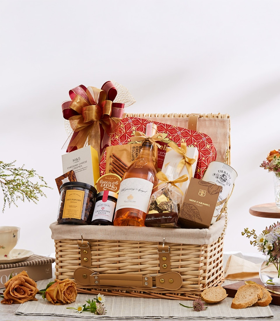 A luxurious wicker basket by Basketeer, brimming with an exquisite selection of gourmet delights: Sauternes wine, decadent chocolates, artisanal jam, gourmet crackers, and delicate biscuits paired with luscious caramel bites. The basket is elegantly adorned with a plush ribbon featuring the Basketeer logo and is artfully surrounded by fresh flowers, artisan bread, and a charming pitcher filled with more blooms—a perfect setting that radiates opulence and inviting warmth.