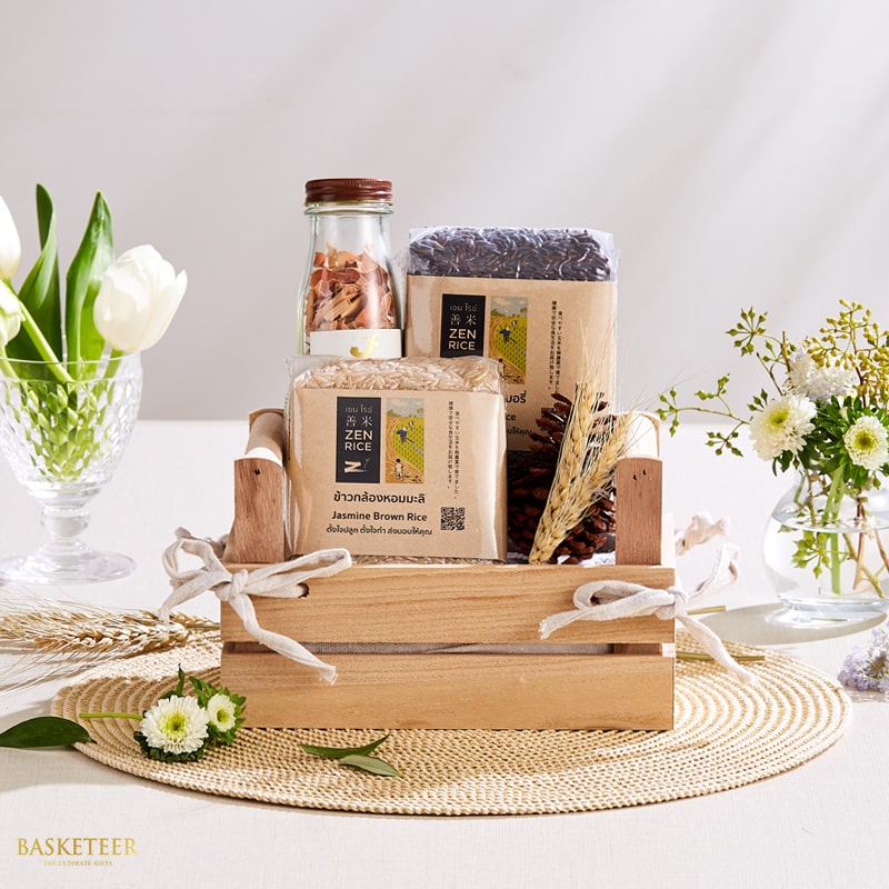 A luxurious wooden basket, beautifully arranged on an intricate woven placemat, proudly showcases its treasures. Nestled within are two varieties of gourmet rice encased in elegant clear packaging, a jar of premium nuts, and a charming pine cone accent. The basket is exquisitely adorned with a pristine white ribbon featuring the Basketeer logo. In the background, delicate white flowers grace a glass vase and a softly glowing candle casts an inviting ambiance. The entire scene exudes sophistication and elegance.
