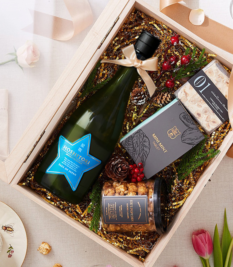 A beautifully curated gourmet gift box featuring a bottle of Hoshitori Japanese sparkling wine, premium caramel popcorn, artisan nougat, and mini mint dark chocolate in an elegant wooden box with a clear lid and gold ribbon.