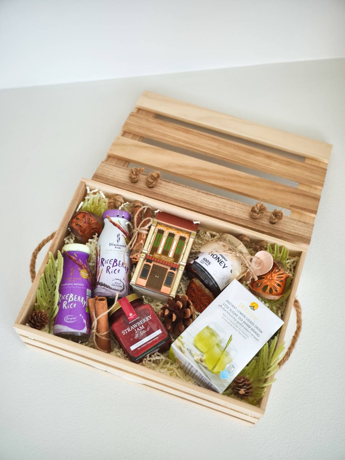 Natural wooden gift box containing riceberry rice, strawberry jam, honey, herbal drink, and assorted gourmet treats with rustic decorations.