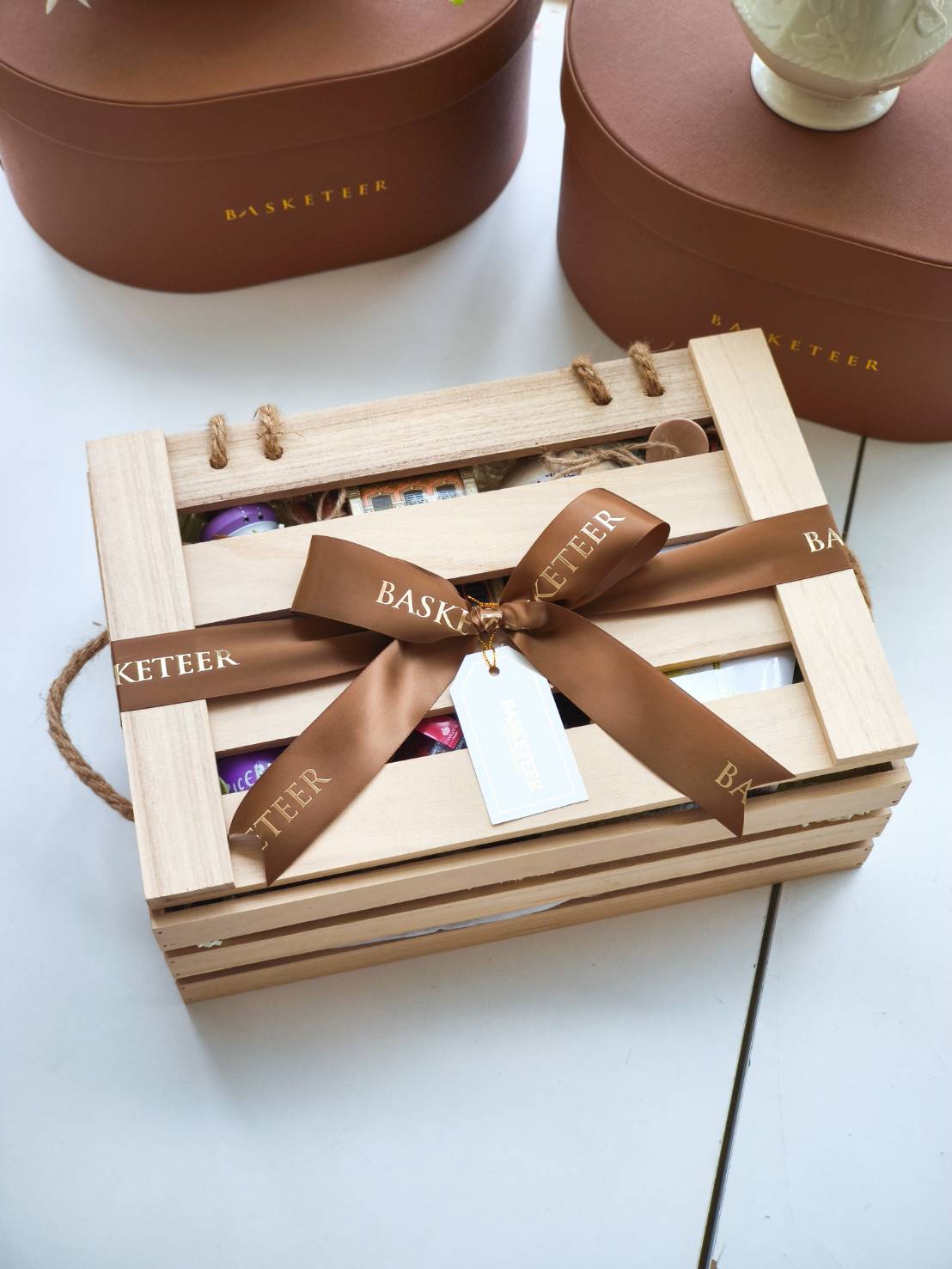 Natural wooden gift box with a brown Basketeer ribbon and gift tag, containing assorted gourmet treats inside.