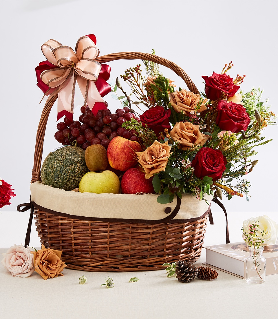 A luxurious wicker basket overflowing with an opulent selection of fresh fruits, including crisp apples, succulent pears, luscious grapes, and juicy melons. This exquisite arrangement is complemented by a stunning display of vibrant red and delicate peach roses interspersed with lush greenery. Adorned with a lavish ribbon bow featuring the elegant Basketeer logo, this Fresh Fruits & Floral Delight is further elevated by charming decorative accents tastefully surrounding the basket. The scene exudes sophistication and invites indulgence in the finest quality produce and florals curated by Basketeer.