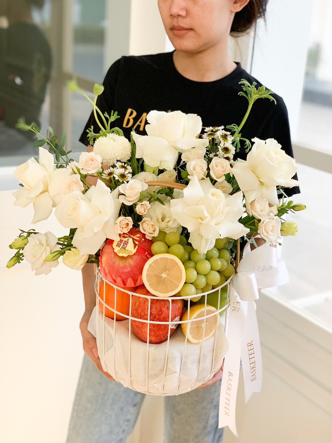 A person holding a white metal basket filled with white flowers, green grapes, two pomegranates, a lemon slice, and greenery. The fresh fruits arrangement is wrapped in a beige cloth and tied with a white ribbon. The person is wearing a black shirt and blue jeans.