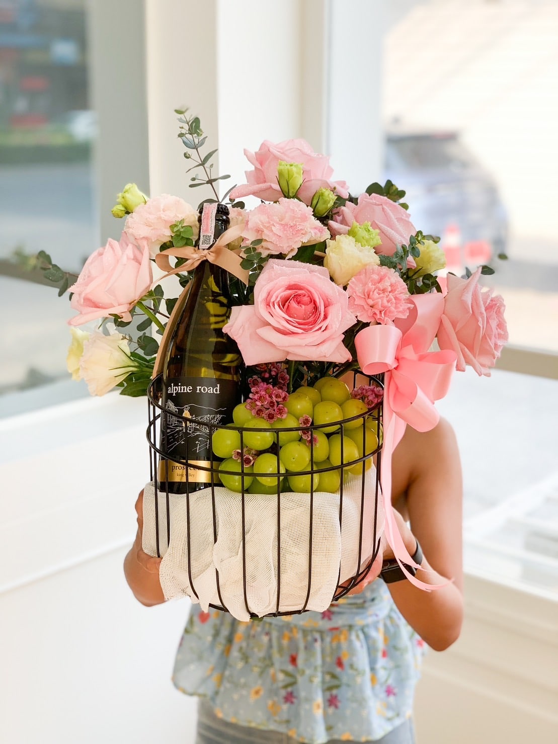 A person holds a gift basket filled with a bouquet of pink roses and carnations, a bottle of wine, and green grapes. The basket is adorned with a pink bow and ribbon, and the person is standing indoors near a window.