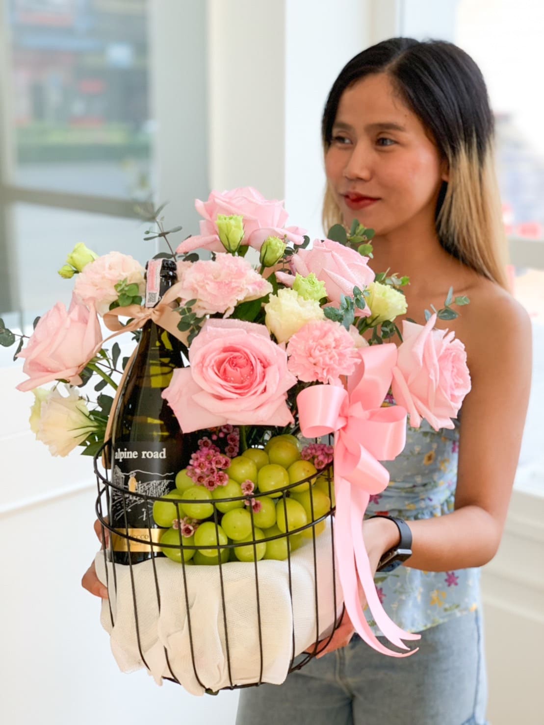 A woman with long, dark blond hair holds a wire basket filled with pink roses, pink carnations, green grapes, and a bottle of wine. The basket is adorned with a large, pink ribbon. She is wearing a light blue, floral sleeveless top and light jeans.