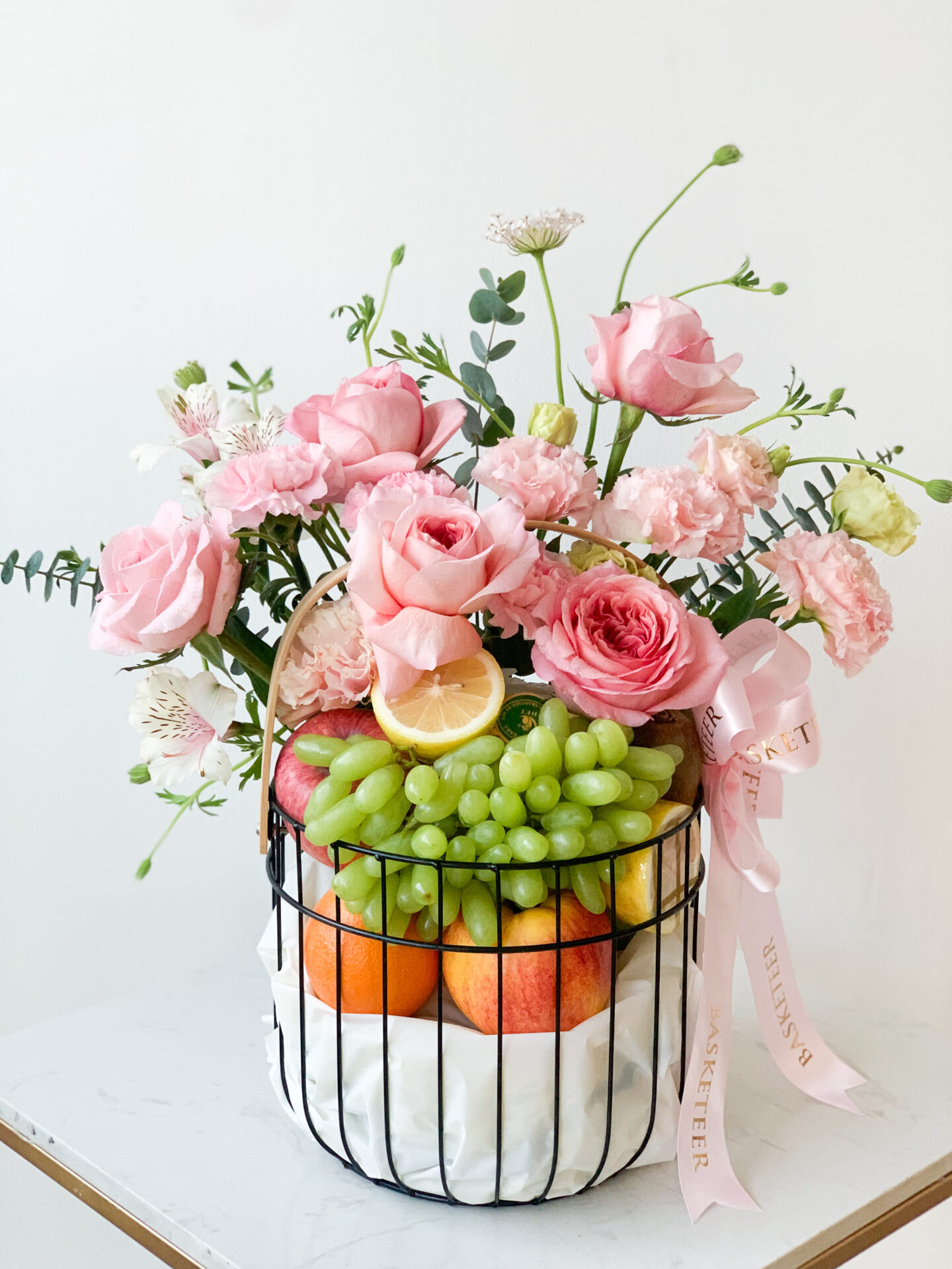 A Sweet Roses Fresh Fruits Basket filled with green grapes, apples, and oranges is adorned with pink roses, carnations, and greenery. A sliced lemon is placed among the roses. A light pink satin ribbon with text is tied to the basket handle. The basket sits on a marbled surface.