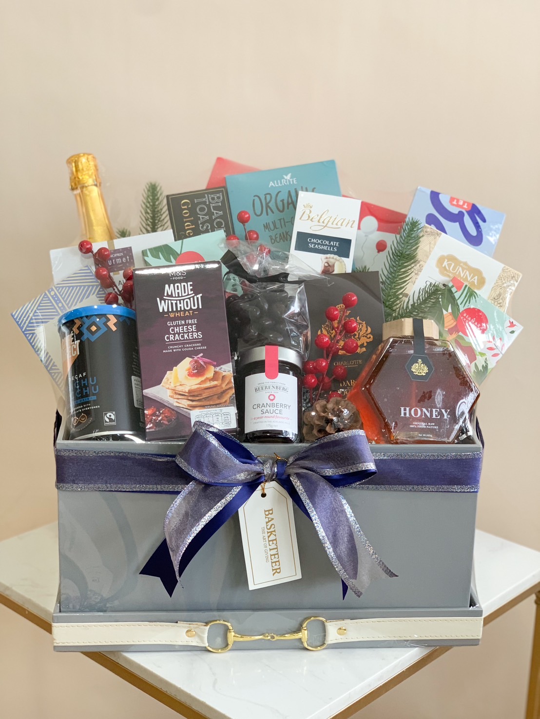 An exquisite Basketeer luxury gift hamper elegantly showcases an array of indulgent treats, including gourmet chocolates, artisanal cookies, premium coffee, and a bottle of sparkling wine. The basket is adorned with a rich purple ribbon bearing the Basketeer logo. Surrounding the hamper are vibrant spring flowers like daffodils and tulips, enhancing the allure. Carefully arranged cookies and crackers lie nearby, completing this scene of refined opulence and inviting warmth.