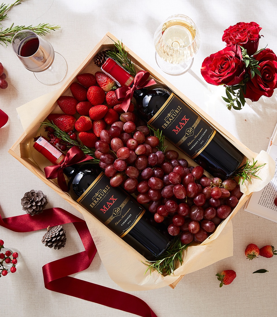 A sumptuous wooden gift box by Basketeer, elegantly adorned with luxurious red ribbons, holds two exquisite bottles of red wine nestled among fresh, plump bunches of grapes, strawberries bursting with flavor, and aromatic rosemary sprigs. The sophisticated scene is completed with a delicate wine glass, beautiful roses in full bloom, rustic pine cones, and scattered strawberries on a pristine white surface. This understated opulence reflects the signature charm and timeless elegance of the Basketeer brand.