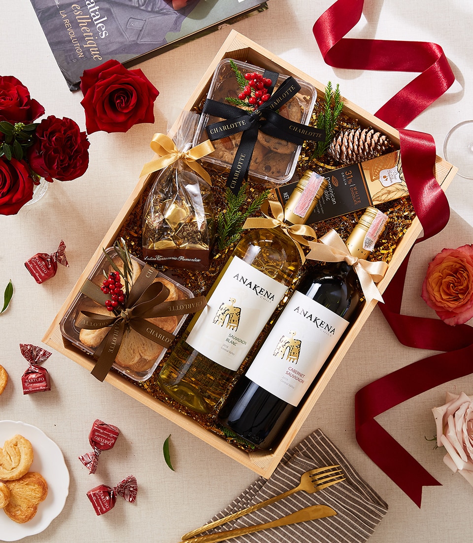 A sumptuous gift hamper from Basketeer featuring exquisite red and white wine bottles, a selection of cookies in sophisticated packaging, decadent chocolates, and vibrant red roses. Adorned with decorative ribbons branded with the Basketeer logo, this luxurious arrangement sits elegantly on a beautifully set table complete with additional chocolates, a glass of fine wine, delicate plates, and ornamental accents. An invitation to indulgence and refined taste.