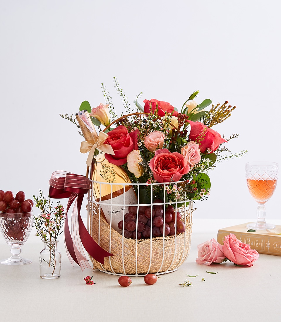 A sumptuous wire basket exudes elegance with a luxurious bottle of champagne, a vibrant bouquet of red and pink roses intertwined with lush green foliage, and an opulent cluster of red grapes. The basket is elegantly adorned with a burgundy ribbon featuring the distinguished Basketeer logo. Nestled beside this lavish arrangement, a glass of exquisite rosé wine awaits, complemented by a delicate vase filled with fresh greens. A dish brimming with perfectly ripened grapes and an inviting book complete the scene, promising a moment of indulgent pleasure curated by Basketeer.