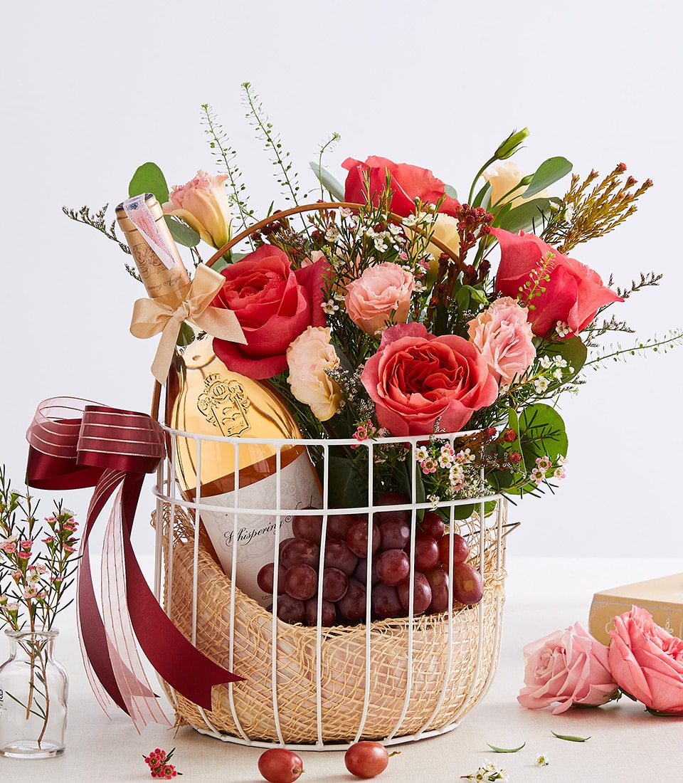A luxurious woven basket with a sleek white metal frame cradles an exquisite arrangement of red and pink roses, complemented by other assorted blooms. Nestled among the flowers is a premium bottle of wine and a succulent bunch of grapes. The basket is elegantly adorned with a sumptuous maroon ribbon, proudly displaying the Basketeer logo. Set against a soft, light background with delicate loose flowers artfully scattered nearby, this scene exudes sophistication and invites indulgence.