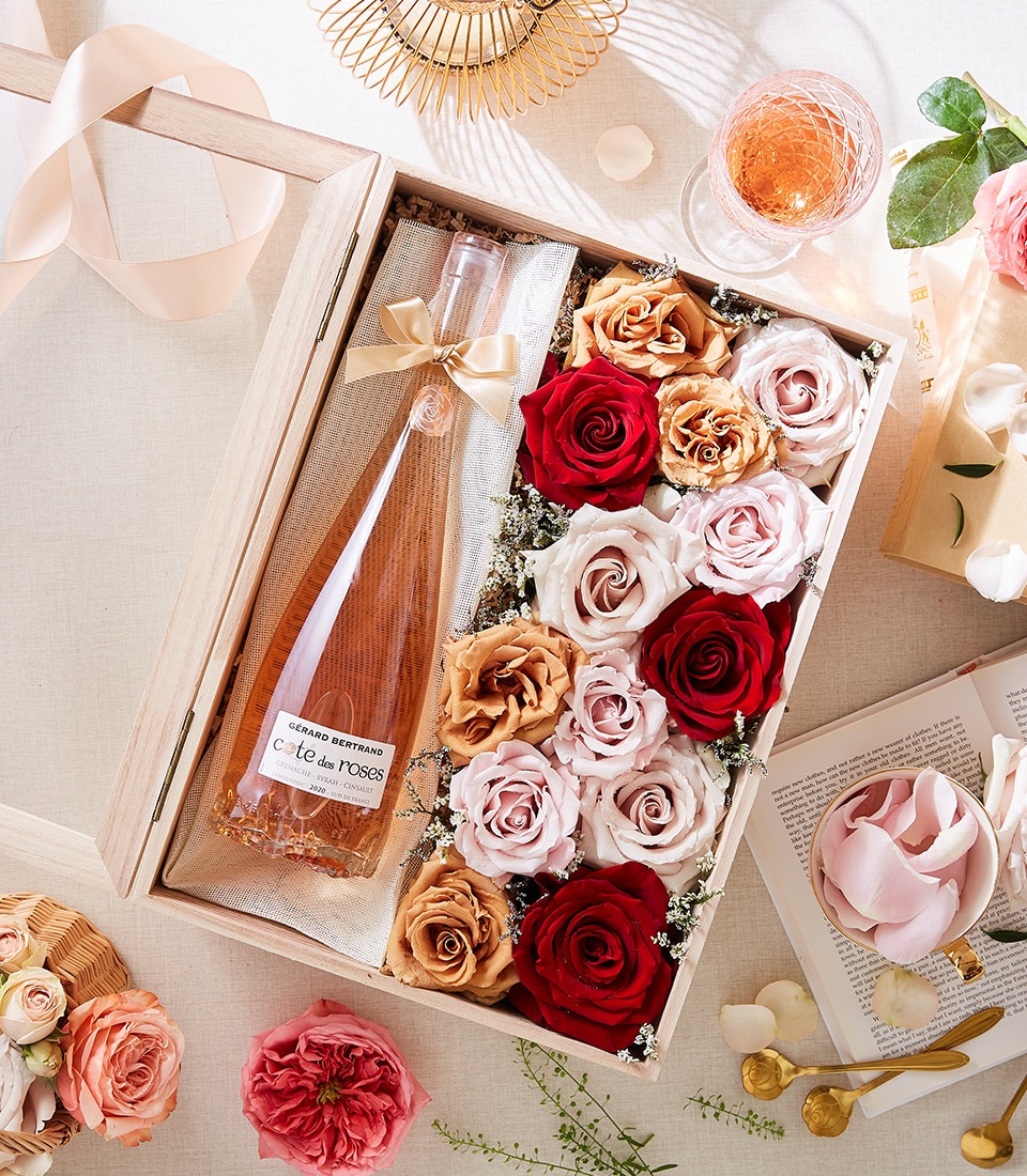 A luxurious wooden box elegantly branded with the Basketeer logo cradles a bottle of exquisite rosé wine surrounded by an assortment of opulent roses in passionate shades of red, blush pink, and creamy beige. The scene is artfully arranged on a polished, sophisticated surface adorned with additional roses, a gracefully presented glass of rosé, an inviting open book, and a delicate silken ribbon bearing the Basketeer name. This Rosé Wine & Romantic Roses Set exudes timeless charm and elevated sophistication.