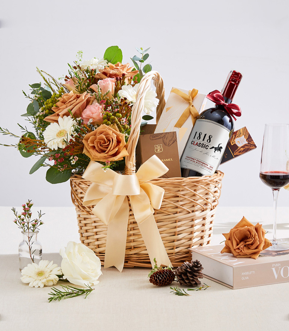 A premium gift basket featuring a bottle of red wine, gourmet chocolates, and an elegant floral arrangement with warm-toned roses and fresh blooms, presented in a woven basket with a gold ribbon—perfect for celebrations, corporate gifts, and special occasions.