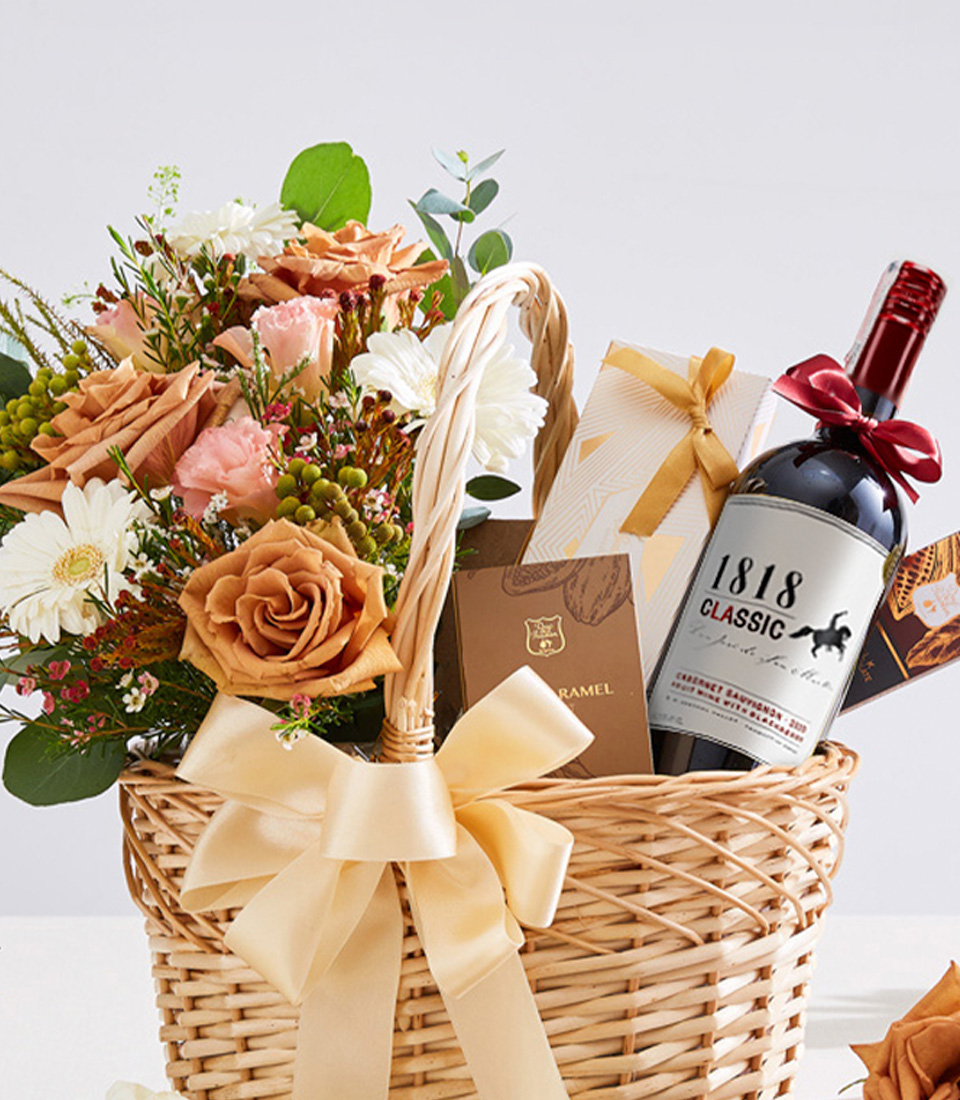 Close-up of a premium gift basket featuring a bottle of red wine, gourmet chocolates, and an elegant floral arrangement with warm-toned roses and fresh blooms, presented in a woven basket with a gold ribbon—perfect for celebrations, corporate gifts, and special occasions.