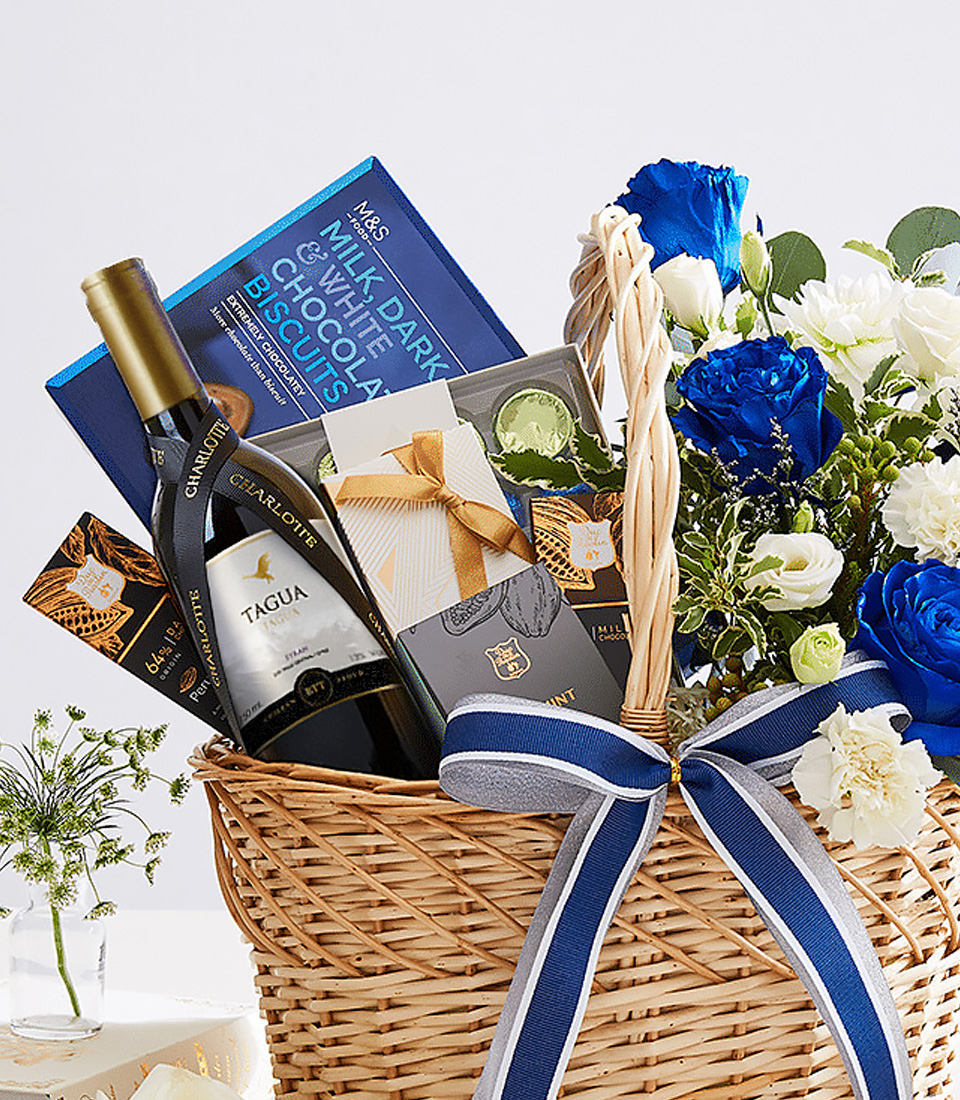 Close-up of a luxury gift basket featuring a bottle of fine white wine, gourmet chocolates, and premium biscuits, elegantly arranged with vibrant blue and white flowers in a woven basket adorned with a navy blue ribbon—perfect for corporate gifting, anniversaries, and celebrations.