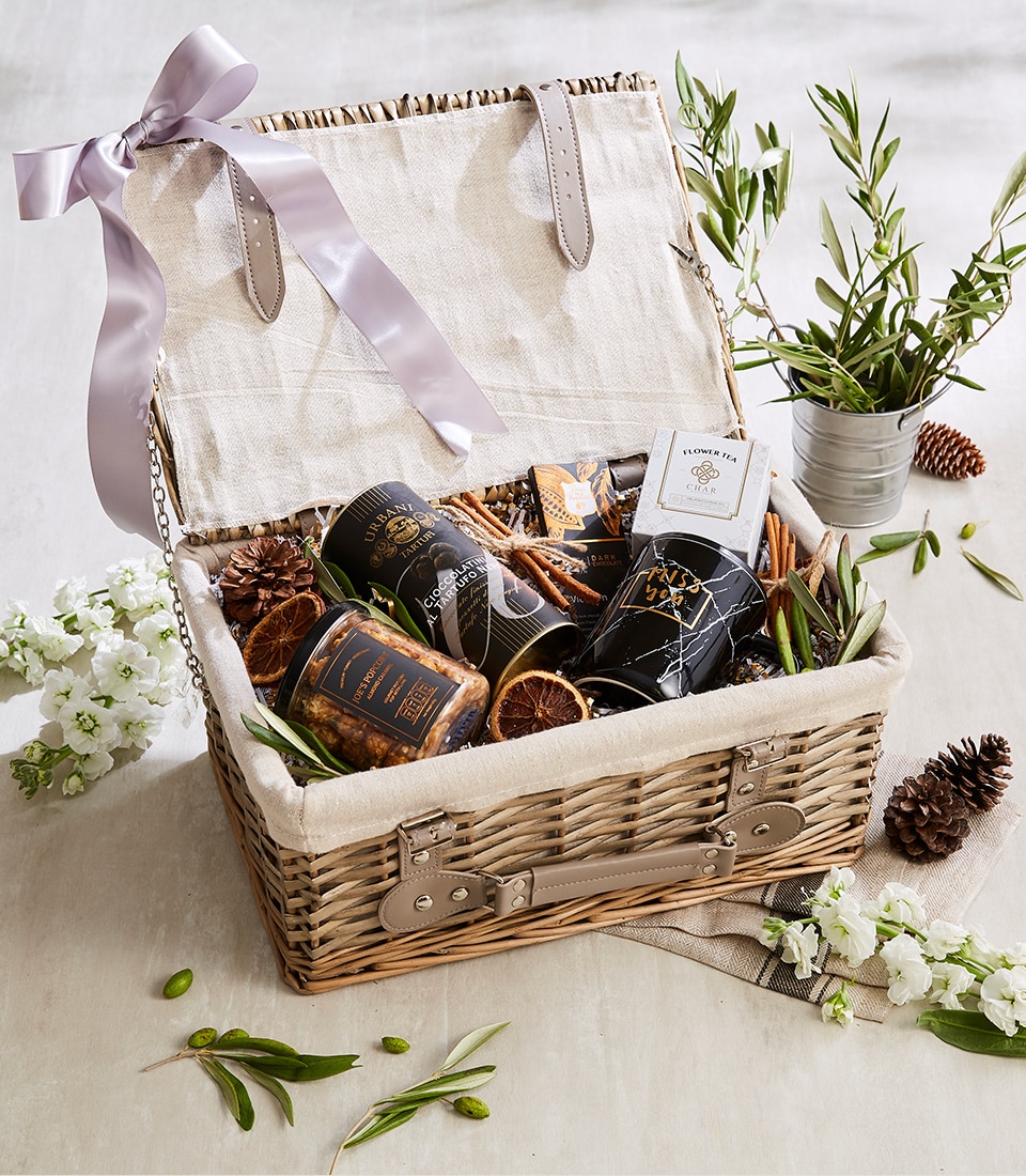 A luxurious wicker gift basket from Basketeer is beautifully open, revealing an exquisite selection of gourmet delights. Inside, elegantly arranged jars, a meticulously crafted small box bearing the Basketeer logo, and artfully dried citrus slices captivate the senses. The basket is gracefully adorned with a light purple ribbon that effortlessly complements its sophisticated presentation. Surrounding the basket are fresh greenery and pine cones, creating a warm and inviting ambiance perfect for any occasion.