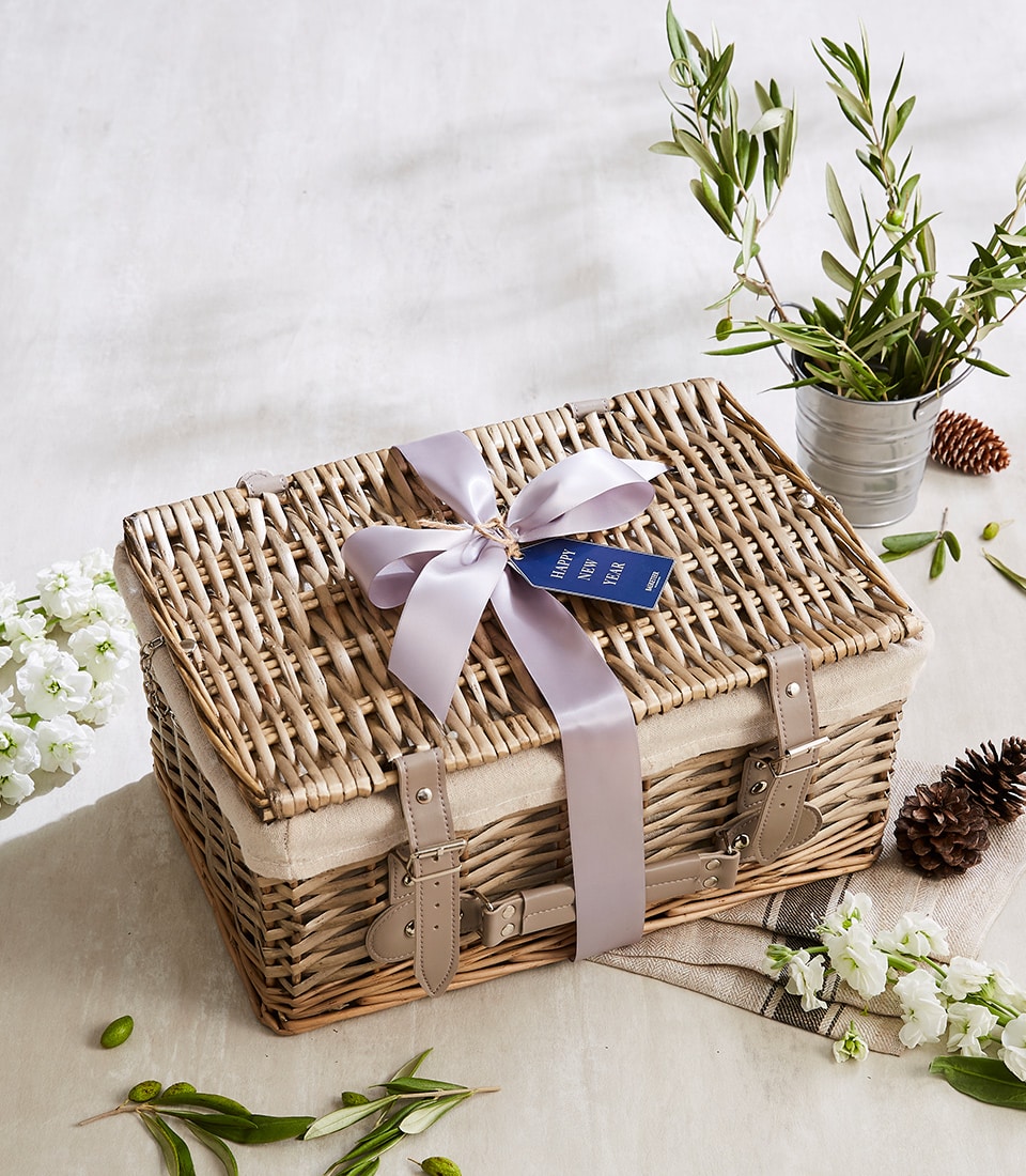 A luxurious wicker gift basket elegantly adorned with a soft lavender ribbon and a refined blue tag, emblematic of Basketeer’s signature touch. Nestled on a light-hued surface, the scene is enriched by delicate white flowers, lush greenery, and an exquisite potted plant, creating an inviting and sophisticated presentation. The Basketeer logo graces the arrangement, ensuring a distinguished allure that captivates the senses.