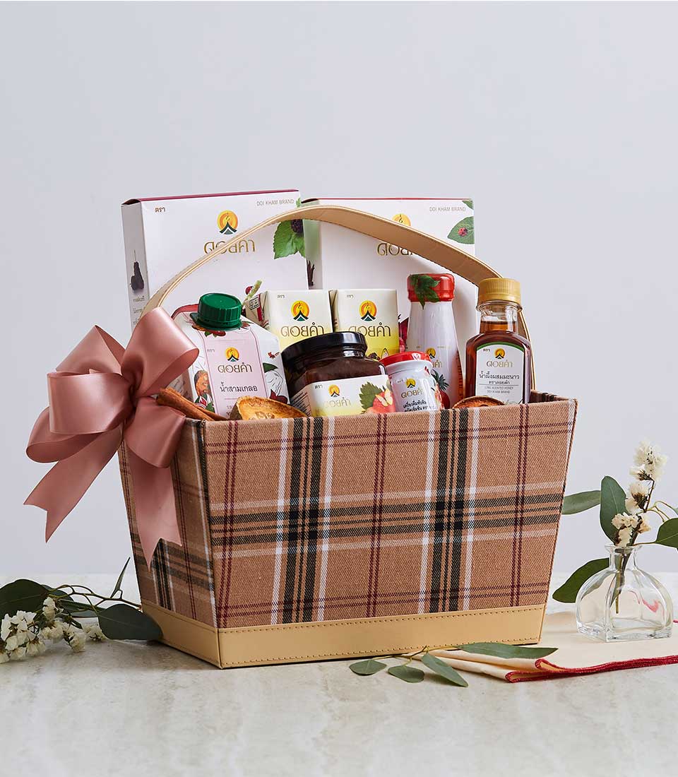A plaid-patterned gift basket with a beige handle and pink bow, resembling the elegant Thai Doi Kham Luxury Bag, contains various food items such as packaged snacks, jars, and bottles. The basket is placed on a white surface with decorative flowers around it.
