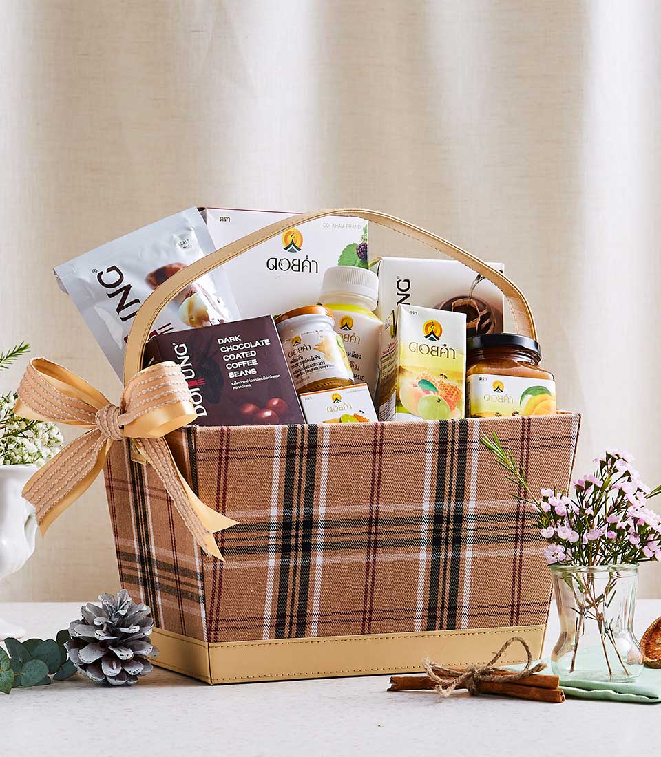 A plaid-patterned gift basket with a cream-colored bow is filled with various packaged items, including snacks, a drink, and other goodies. The basket is placed on a white surface with pine cones and small pink flowers nearby.