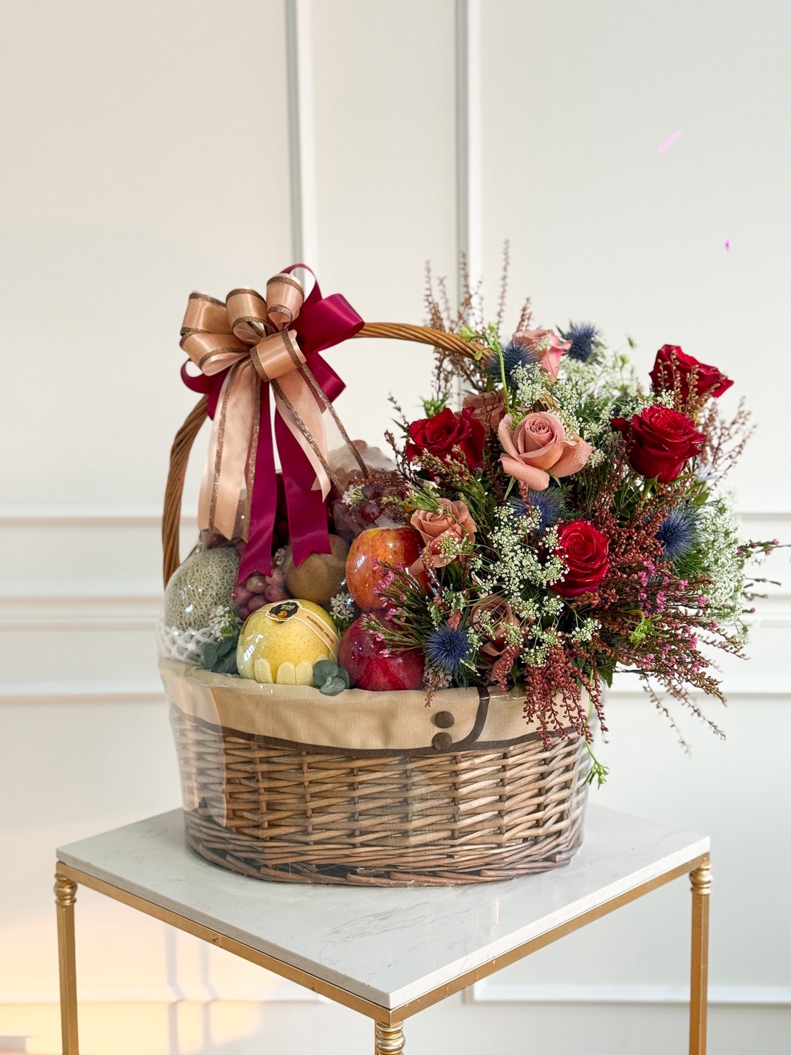 A wicker basket on a table, filled with Fresh Fruit & Floral Delight—assorted fruit adorned with red and pink roses, greenery, and a large gold and maroon ribbon bow on the handle.