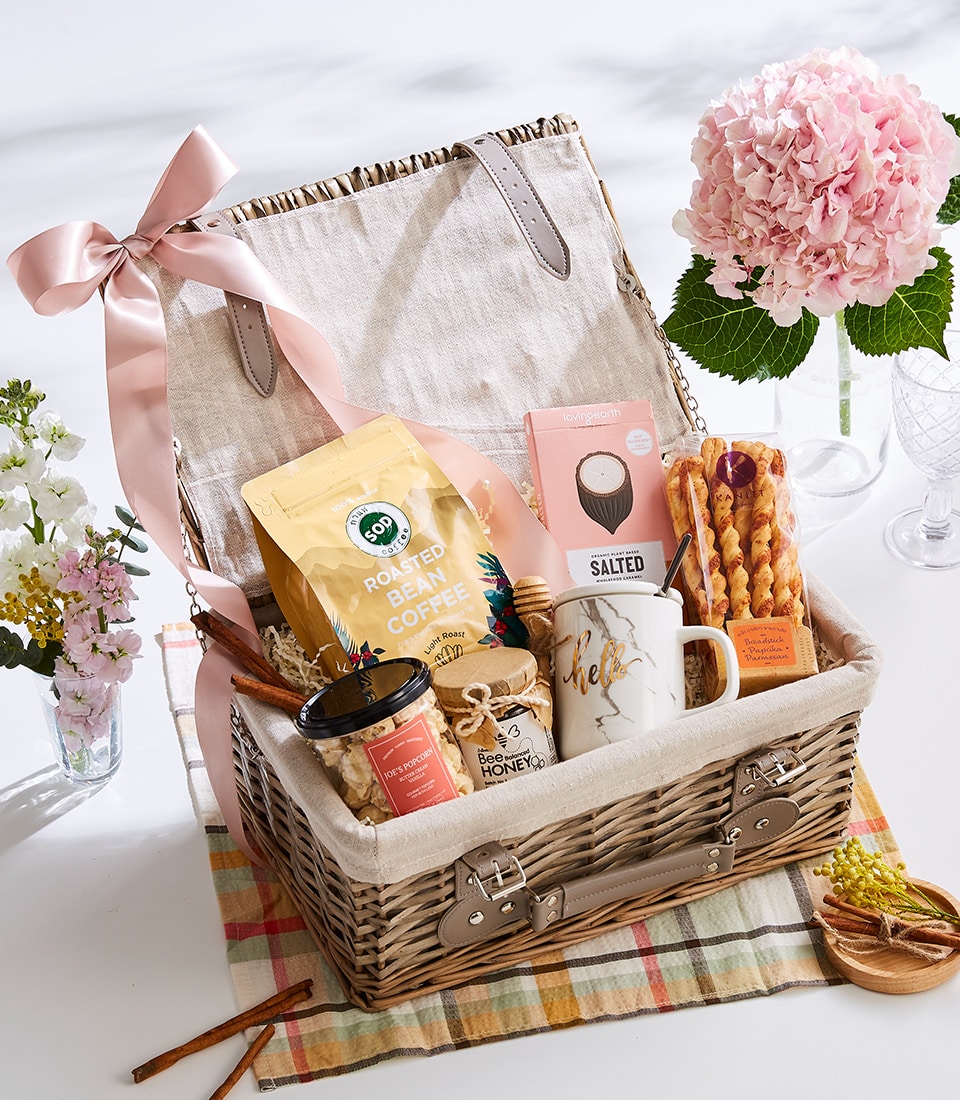 A luxurious wicker gift basket adorned with a delicate pink bow features an exquisite selection of Basketeer delights. Nestled within are premium roasted coffee beans, an elegantly frosted candle, a jar of golden honey, gourmet salted treats, a finely crafted ceramic mug, and decadent chocolate. The basket sits on an inviting plaid cloth, surrounded by fresh flowers arranged in glass vases on a pristine white surface. The enchanting scene radiates warmth and sophistication, highlighted by the Basketeer logo subtly displayed on a ribbon enhancing the overall allure.