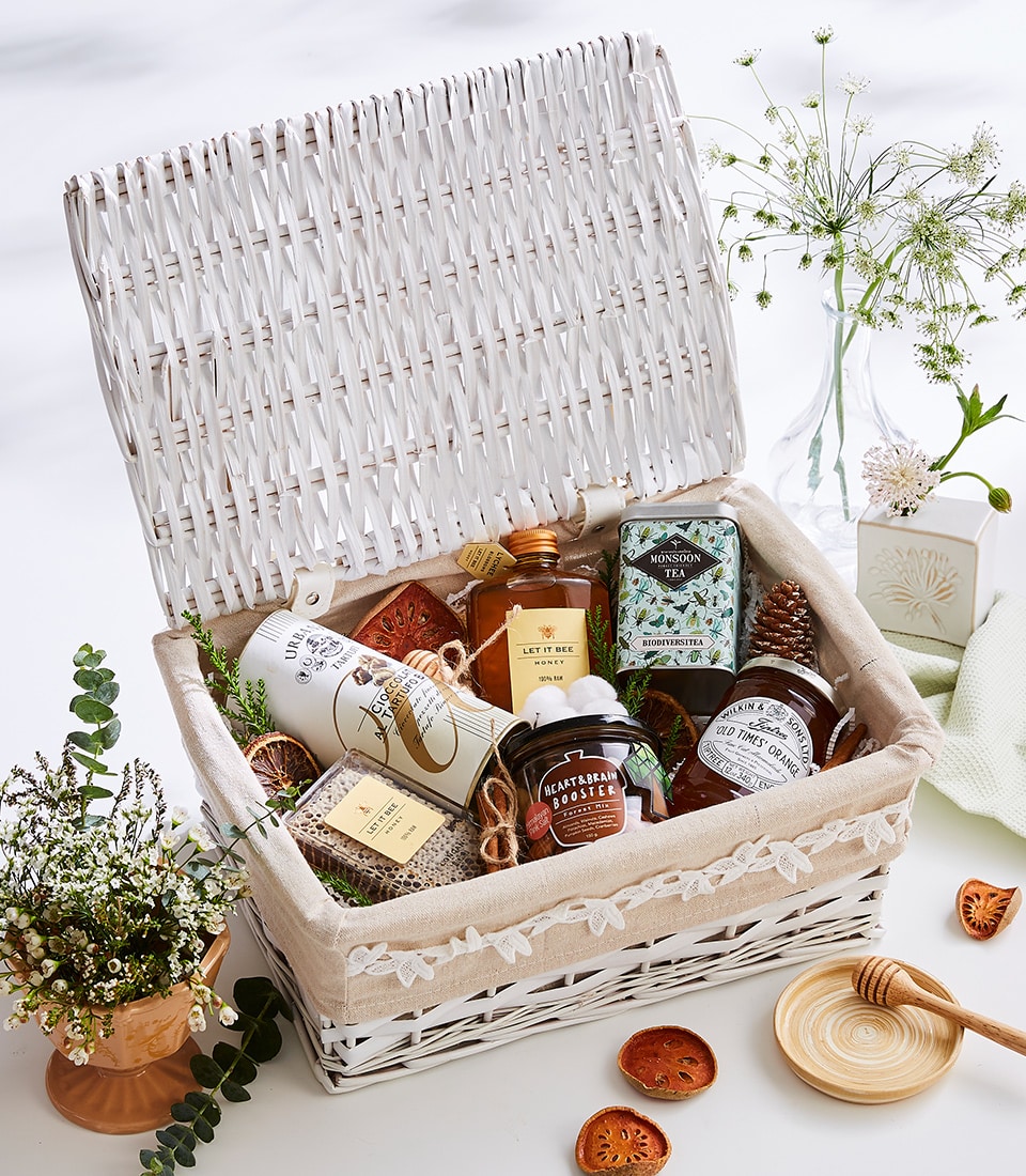 A luxurious white woven basket, meticulously curated by Basketeer, brimming with an exquisite selection of gifts. This elegant ensemble includes artisanal jars of honey, a premium tin of tea, an indulgent bottle of lotion, and an array of other thoughtfully packaged goods. The basket is tastefully lined with soft fabric and surrounded by charming accents such as delicate small plants, a handcrafted honey dipper, and aromatic dried orange slices. A ribbon bearing the Basketeer logo adds a touch of sophistication to this enticing scene.