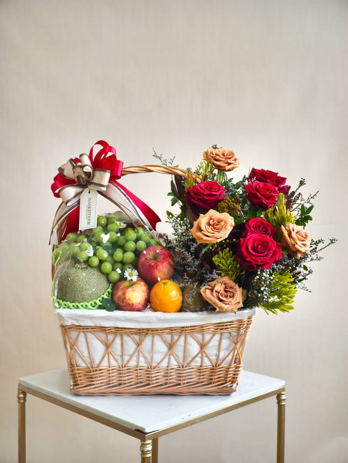 Wicker gift basket with fresh fruits and mixed roses in red and peach tones, tied with red and champagne ribbon