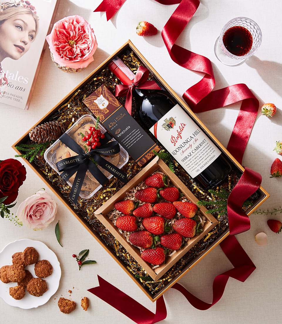 A lavishly curated Basketeer gift box exudes festive elegance, showcasing a premium bottle of red wine nestled alongside a succulent box of strawberries. Delight in the assorted gourmet confections and the exquisitely adorned charcuterie package, each item presented to perfection. The scene is accentuated by luxurious red ribbons bearing the Basketeer logo, delicately arranged flowers, and an inviting scattering of cookies and rose petals across the table.