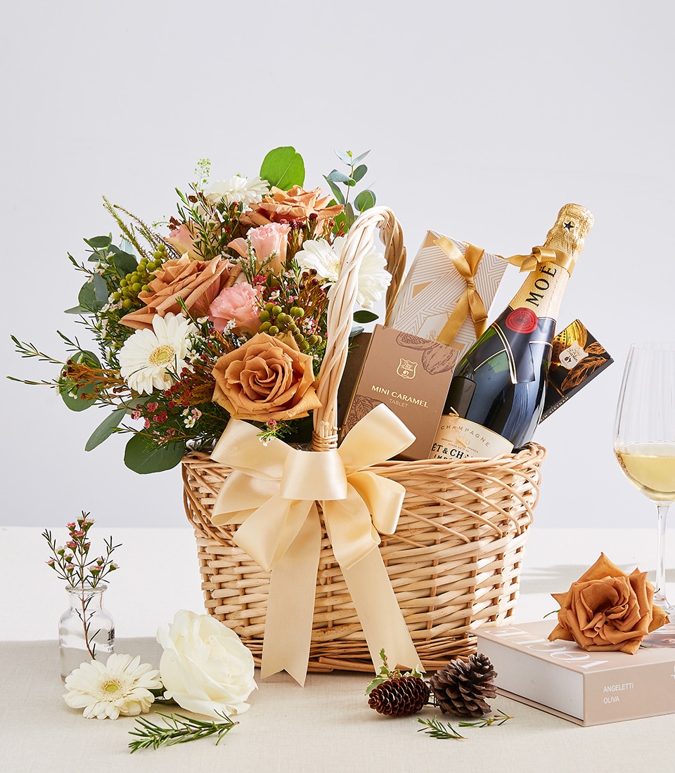 A luxurious wicker basket featuring an exquisite bouquet of flowers, a prestigious bottle of Moët & Chandon champagne, and an assortment of gourmet delicacies and elegant gifts from Basketeer. The arrangement is gracefully adorned with a sumptuous gold ribbon bearing the Basketeer logo, and is elegantly presented on a light-colored tablecloth surrounded by additional delicate flowers and charming pine cones.