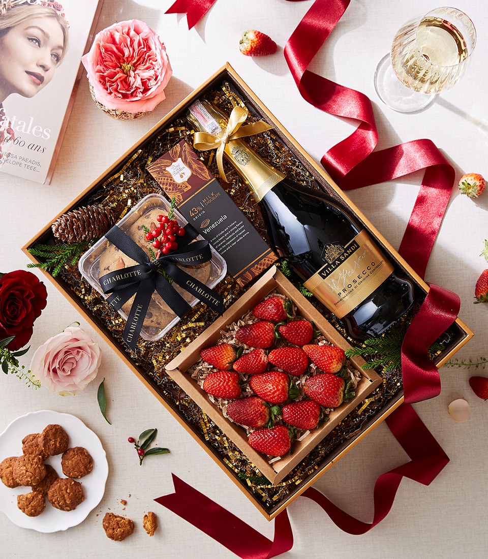 An opulent gift box from Basketeer, adorned with their signature logo, showcases a meticulously curated selection of indulgences: a bottle of premium prosecco, luscious strawberries, decadent chocolates, and gourmet nuts. Surrounding this luxurious ensemble is an elegant red ribbon branded with the Basketeer name, delicate flowers in full bloom, a glass filled with sparkling prosecco ready to be savored, and an inviting book promising moments of relaxation. Scattered strawberry leaves and petals embellish the scene further, augmenting the festive ambiance and sophisticated presentation.
