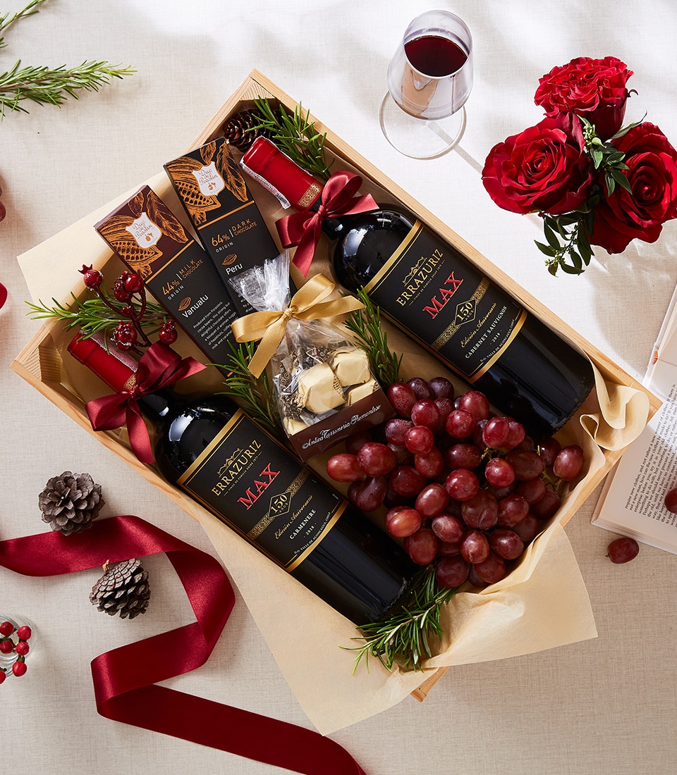 A sumptuous wooden gift tray by Basketeer presents itself with two exquisite bottles of red wine, surrounded by an array of rich dark chocolate bars and a cluster of plump, juicy red grapes. A delicate bag of white chocolates, elegantly tied with a ribbon bearing the Basketeer logo, nestles amidst evergreen sprigs. The scene is enhanced by a filled glass of velvety red wine, complemented beautifully by vibrant red roses and rustic pine cones, evoking undeniable luxury and warmth.