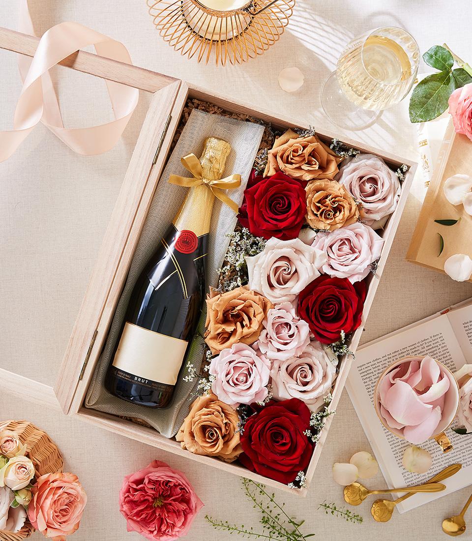 A Luxury Champagne and Flowers Gift Box featuring a wooden container with a bottle of champagne adorned with a gold ribbon, nestled beside an array of roses in shades of red, pink, and peach. The box is surrounded by scattered rose petals, an open book, a rose, a ribbon, and a glass of champagne.