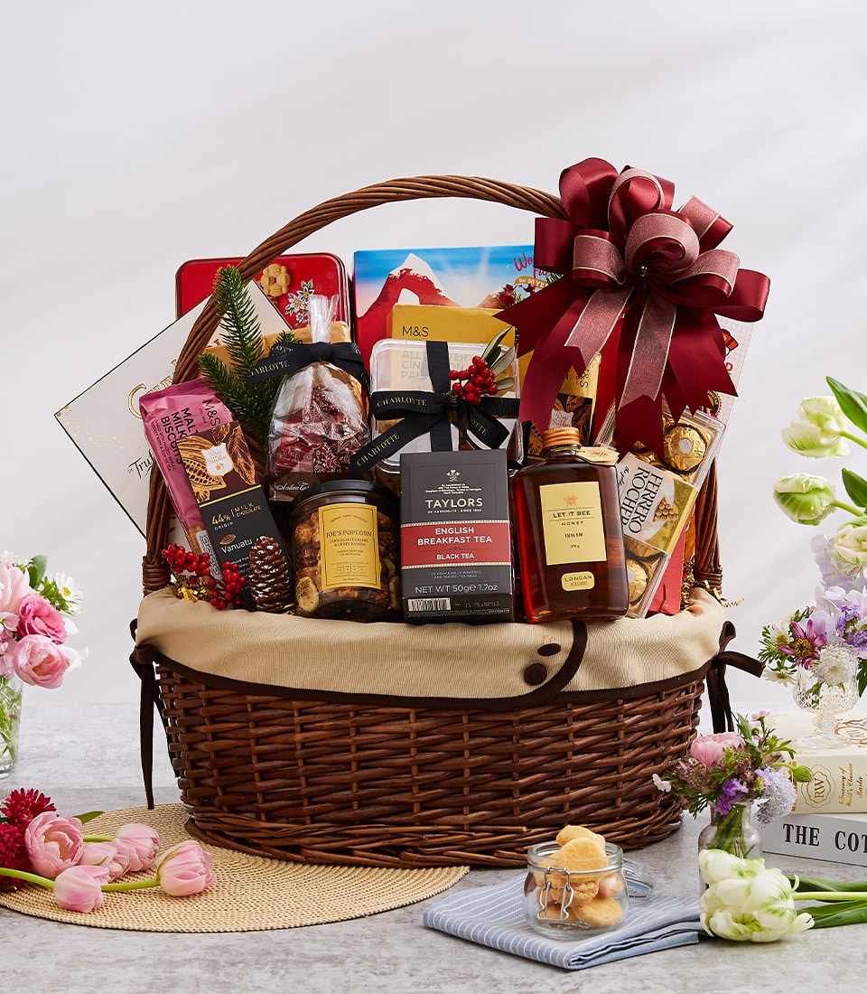 An opulent, large woven gift basket filled with an exquisite selection of gourmet snacks and beverages curated by Basketeer, featuring premium teas, delectable biscuits, sumptuous chocolates, and artisanal jams. The luxurious basket is elegantly adorned with a rich red ribbon bearing the distinguished Basketeer logo, surrounded by fresh flowers on a serene light-colored surface.