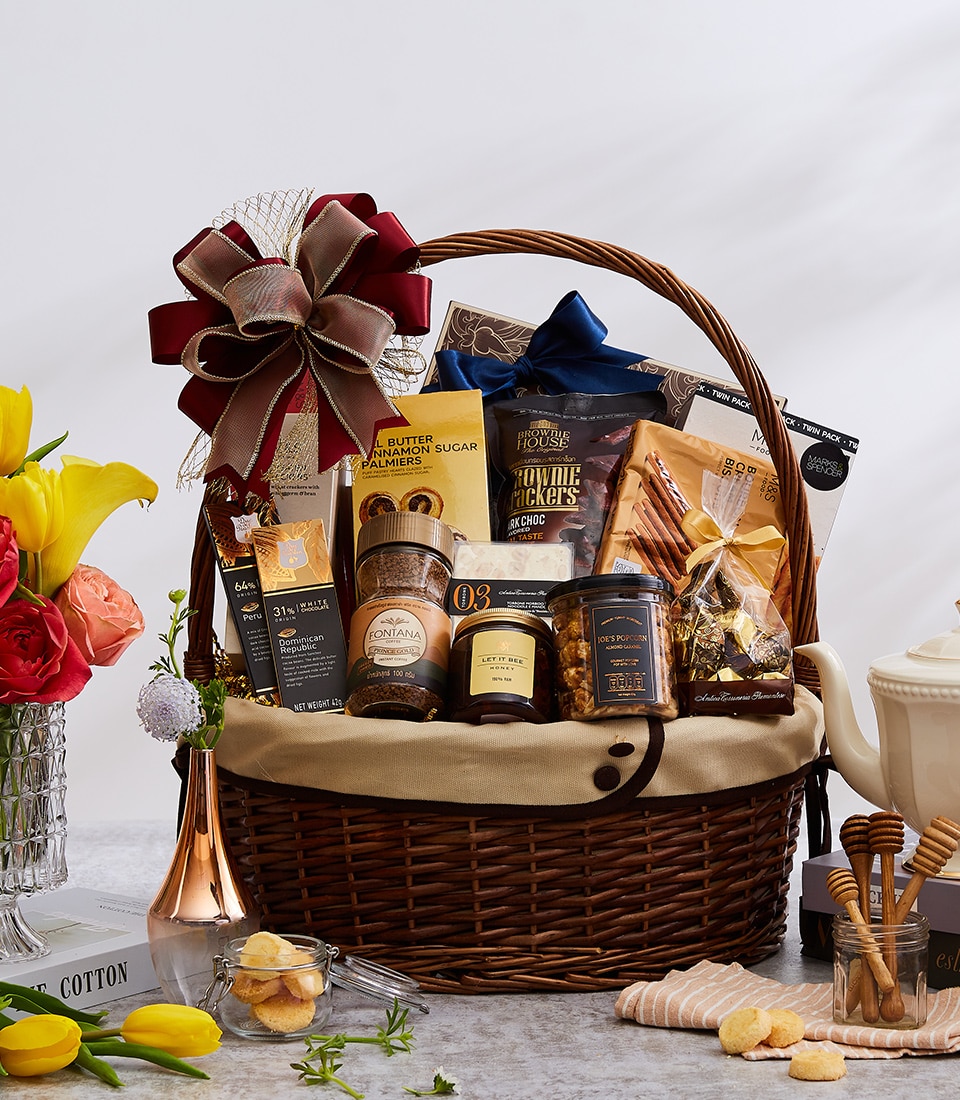 A beautifully crafted wicker gift basket, elegantly adorned with a luxurious red and gold ribbon bearing the Basketeer logo, brims with an exquisite selection of gourmet delights. Nestled within are delectable cookies, savory snacks, rich coffee, and artisanal preserves. This inviting scene is set on a tastefully arranged table featuring a classic white teapot and complemented by vibrant yellow and red flowers along with charming decorative elements that add an extra touch of sophistication.