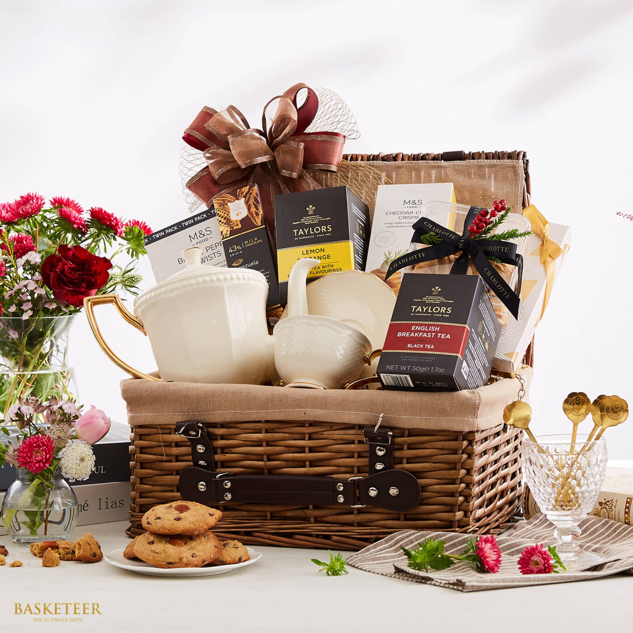 A lavish wicker gift basket filled with premium tea products, accompanied by an elegant white teapot and pitcher, delectable assorted cookies, and a variety of gourmet snacks. The renowned Basketeer brand ribbon adorns the basket, adding an extra touch of sophistication. Surrounding this luxurious ensemble are vases filled with delicate pink and white flowers and a charming plate showcasing more delightful cookies. The scene exudes an inviting and cozy atmosphere perfect for indulgence.