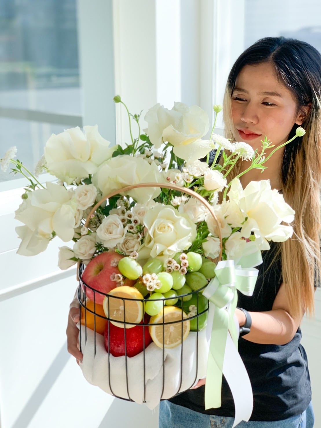 A woman with long hair, wearing a black shirt, holds a sweet roses fresh fruits basket filled with white roses, assorted fruits including apples, grapes, and a lemon. The basket is adorned with a green ribbon and is positioned near a sunlit window.